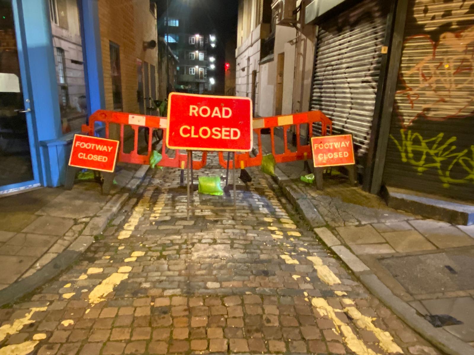 Illuminated road closed and footway closed signage on a cobbled alley at night