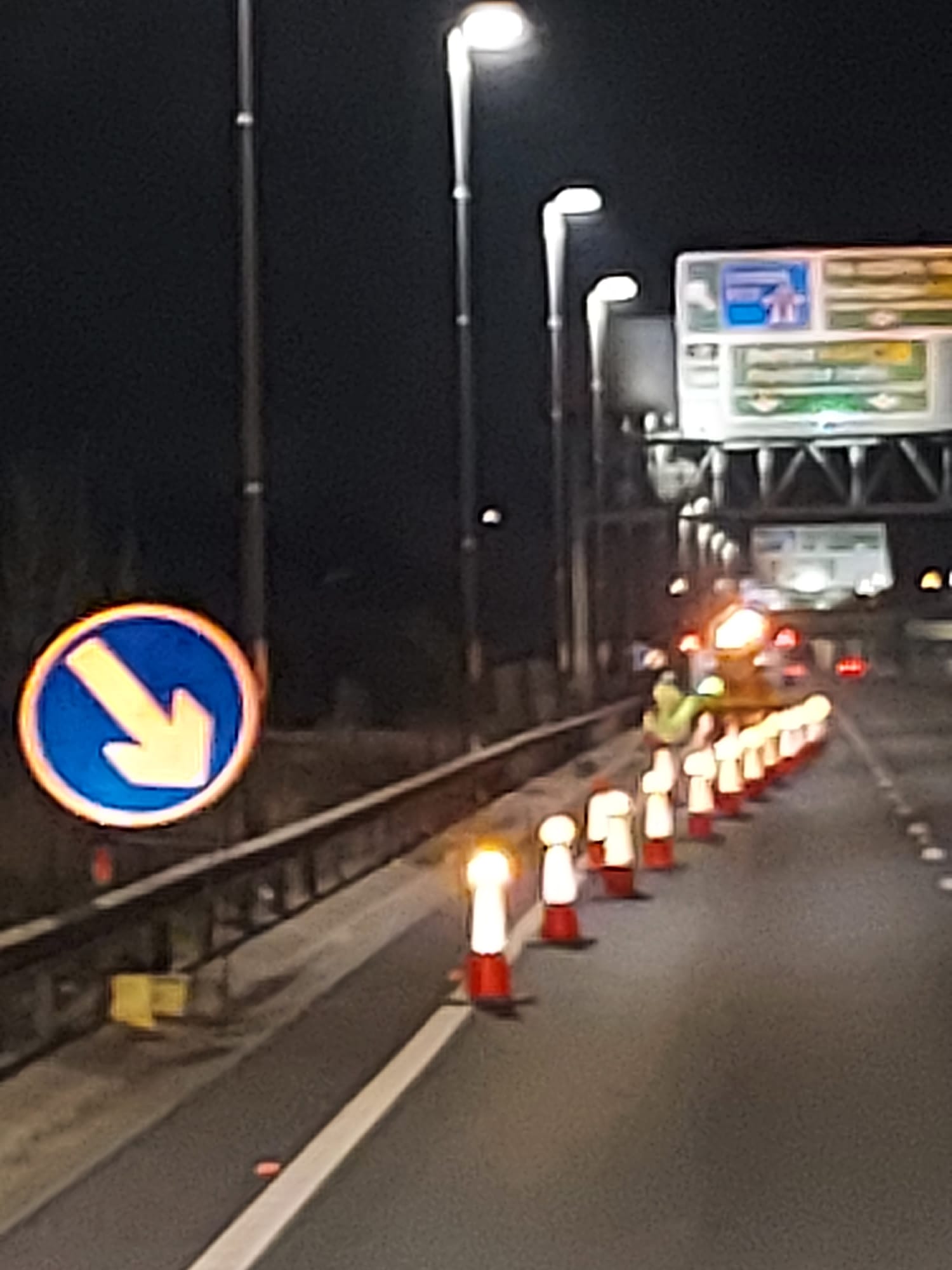 Lane closure arrow sign and cones beneath a motorway gantry at night