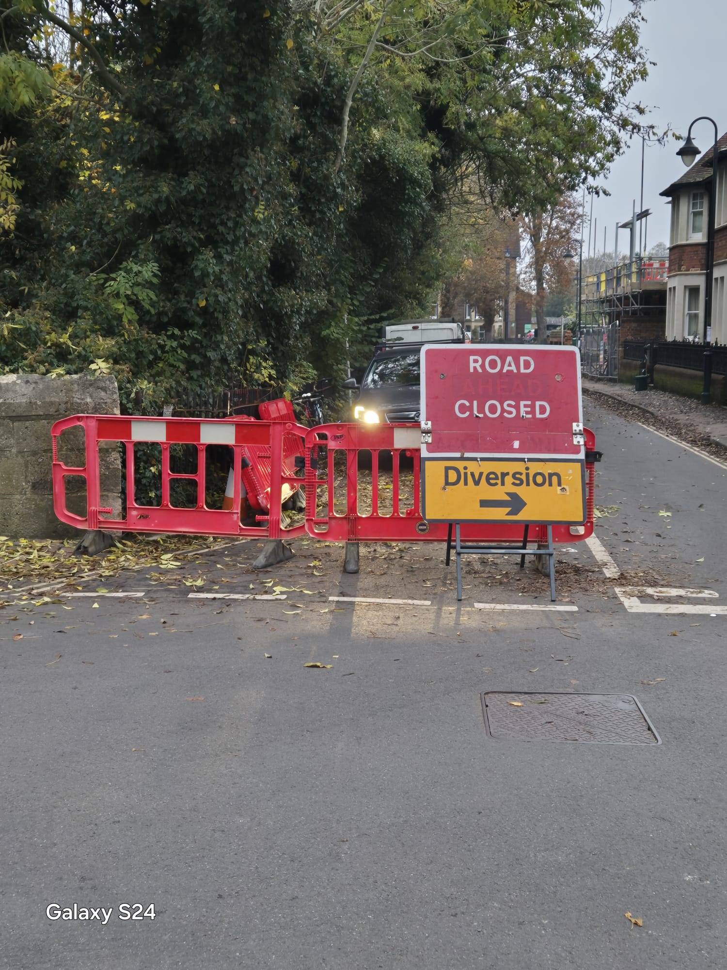 Road closed sign and diversion arrow on barriers closing a tree-lined street at dusk